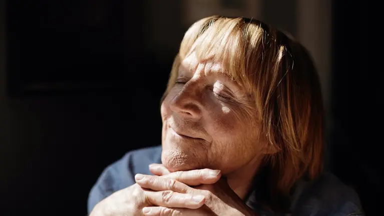 Smiling blond senior woman with hands on chin relaxing in sunlight at home