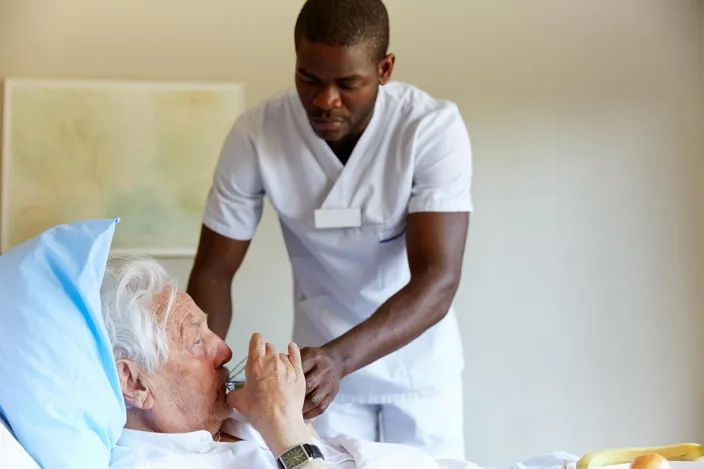 Male nurse feeding water to senior man in hospital ward