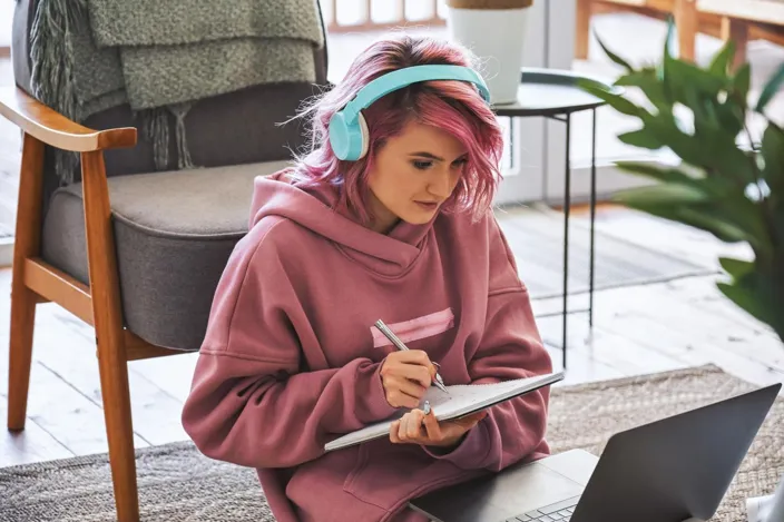 Focused hipster teen girl school college student pink hair wear headphones write notes watching webinar online video conference calling on laptop computer sit on floor working learning online at home.