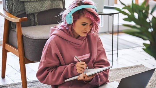 Focused hipster teen girl school college student pink hair wear headphones write notes watching webinar online video conference calling on laptop computer sit on floor working learning online at home.