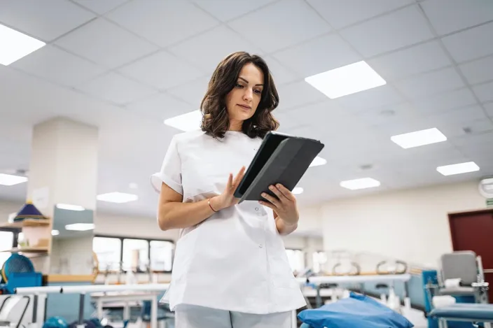 Low angle of brunette in medical uniform browsing data on digital tablet while working in spacious physiotherapy room of modern clinic