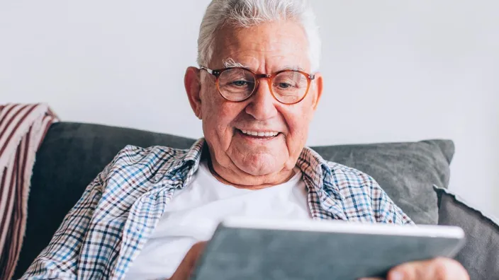 A close up of an old man in his 70s sitting on the couch at home, searching the Internet on his digital tablet, enjoying