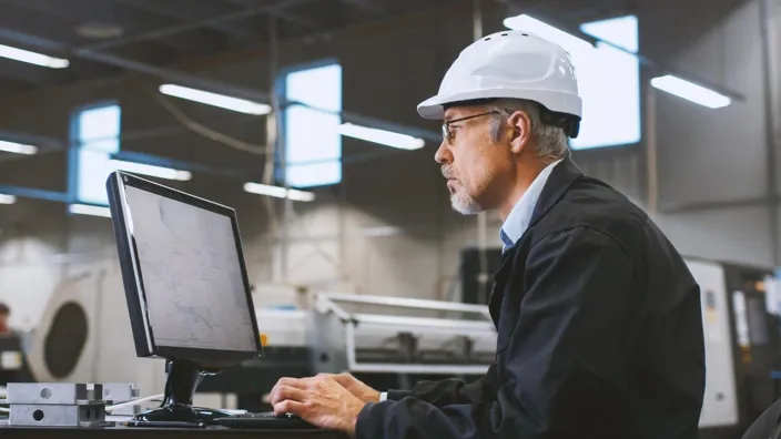 Senior engineer in glasses is working on a desktop computer in a factory.