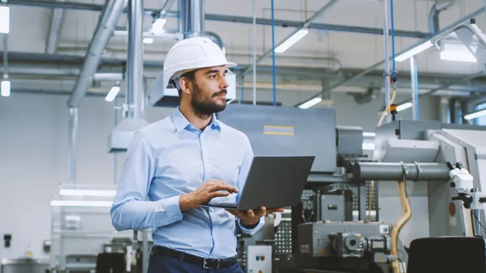 Chief Engineer in the Hard Hat Walks Through Light Modern Factory While Holding Laptop. Successful, Handsome Man in Modern Industrial Environment.