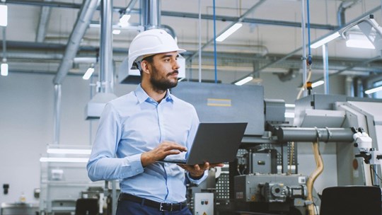 Chief Engineer in the Hard Hat Walks Through Light Modern Factory While Holding Laptop. Successful, Handsome Man in Modern Industrial Environment.