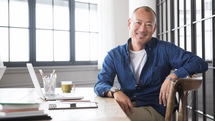 Portrait of mature Japanese smiling man sitting at desk with laptop computer and digital tablet