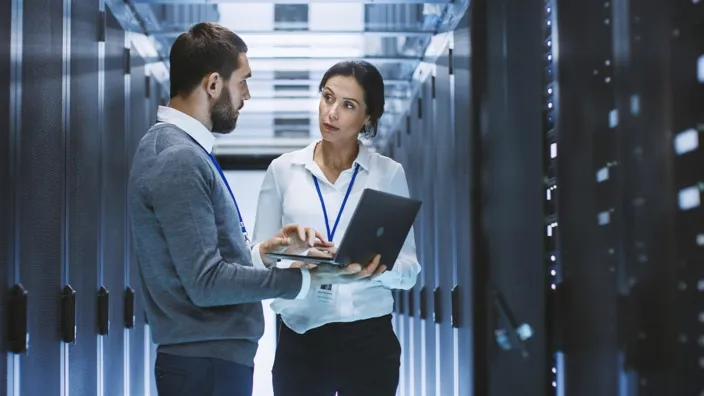 Male IT Specialist Holds Laptop and Discusses Work with Female Server Technician. They're Standing in Data Center, Rack Server Cabinet is Open.