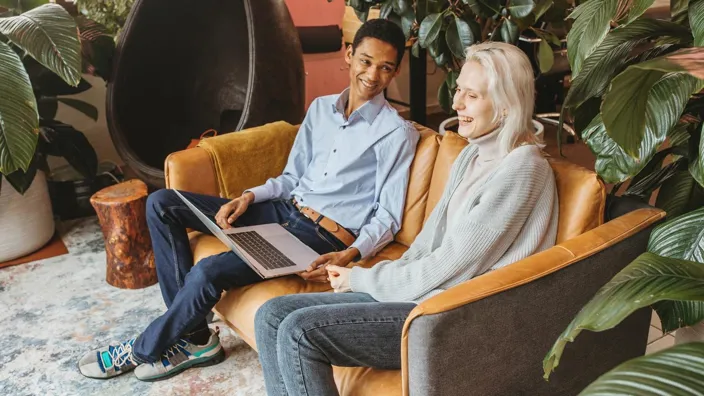 a man and a woman sitting on a sofa, looking at a laptop, laughing