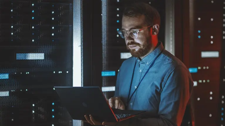 Bearded IT Specialist in Glasses is Working on Laptop in Data Center while Standing Near Server Rack. Running Diagnostics, Doing Maintenance Work. Emergency Red Light from Side Illuminating Specialist
