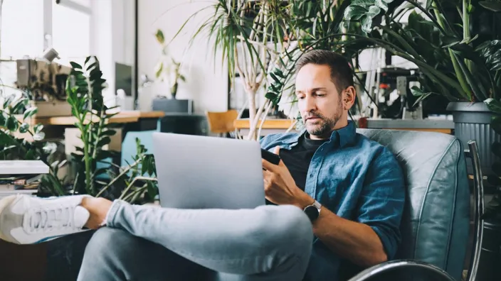 man in armchair with laptop in lap using mobile phone