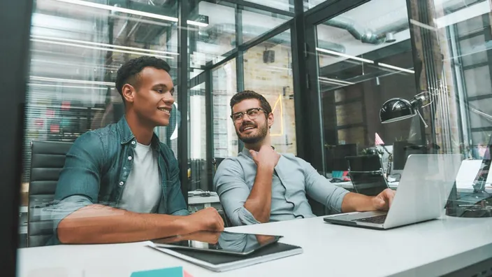 Two young men in meeting in office using laptop and tablet smiling
