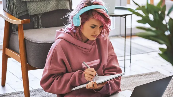 Focused hipster teen girl school college student pink hair wear headphones write notes watching webinar online video conference calling on laptop computer sit on floor working learning online at home.