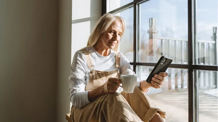 Older woman sitting next to a window checking her smartphone and drinking coffee