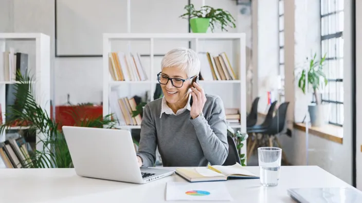 Senior female entrepreneur working in the office and talking on her smartphone, simultaneously checking her laptop