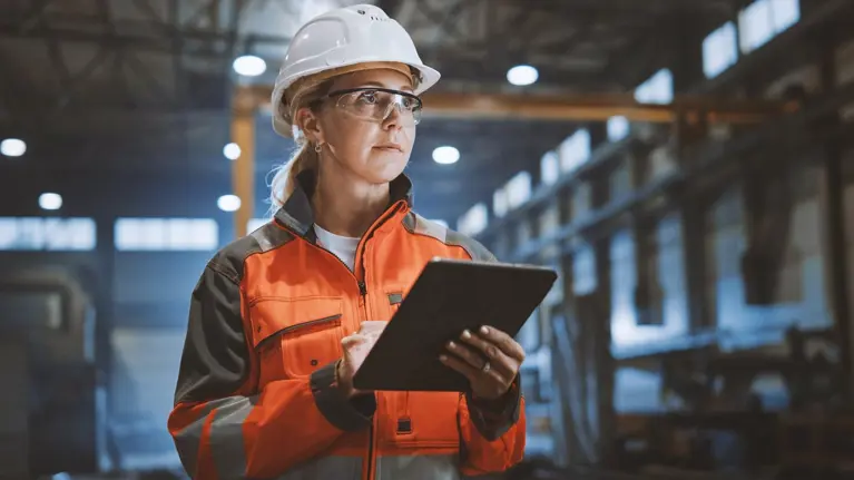 Professional Heavy Industry Engineer Worker Wearing Safety Uniform and Hard Hat Uses Tablet Computer. Serious Successful Female Industrial Specialist Walking in a Metal Manufacture Warehouse.